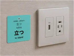 A 立つ (tatsu, to stand) vocabulary sticker on a wall beside an electrical outlet
