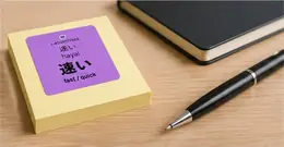 A 速い (hayai, fast/quick) vocabulary sticker on a stack of sticky notes next to a notebook and pen