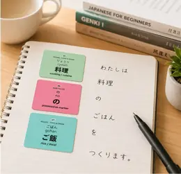 JLPT vocabulary stickers for 料理 (ryouri, cooking), の (no, possessive marker), and ご飯 (gohan, rice/meal) in a notebook next to a stack of Japanese textbooks and a cup of coffee